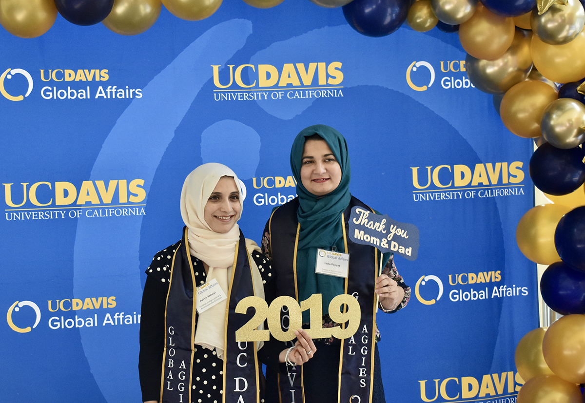 two women in hijab standing in front of a blue background with balloons holding a sign that says 2019