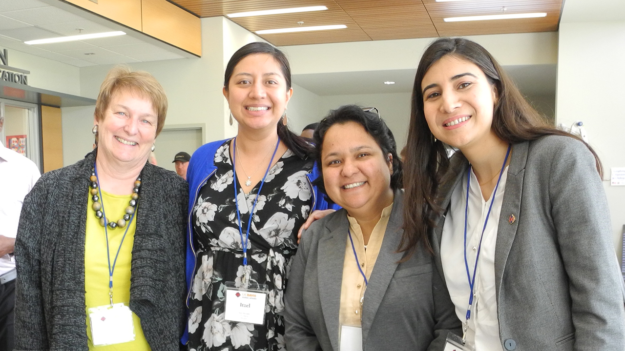 Four women stand inside the International Center, smiling to camera. On the left, Joanna Regulska with short blonde hair wears a yellow blouse under a black and gray knit cardigan and large beaded necklace. Next to her, Itzel Morales has her long dark hair tied back in a low ponytail and wears a black and white floral top under a bright blue jacket. Humera Qasim Khan wears a button-up collarless shirt in cream under a light gray blazer. Her glasses sit atop her short dark hair. On the very right,