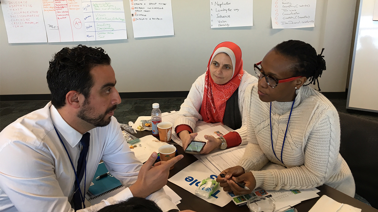 UC Davis Humphrey Fellowship Program Fellows Adnane Labacci wears a white collared shirt and dark tie as he speaks to Maria Rodriguez seated across the table from him and wearing black and red glasses and a white knit sweater. Looking on from Maria’s right is Heba Allah Essam Khalil wearing a white cardigan over a dark top and an orange hajib. Papers are scattered on the table between them, and large sheets of note paper hang on the walls around them. 