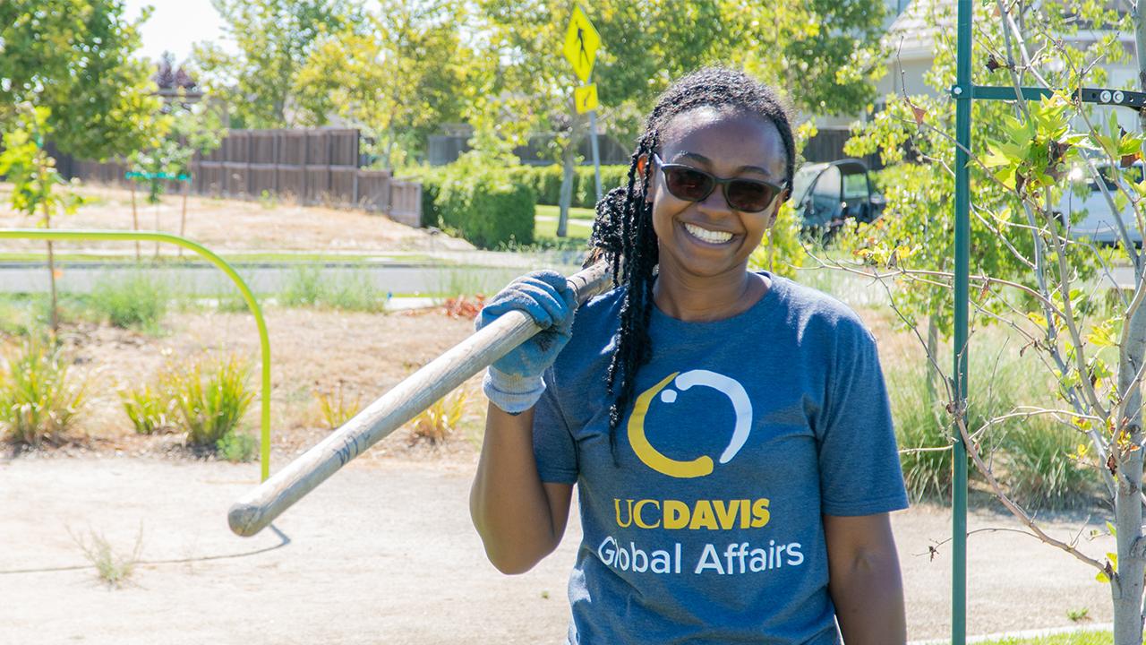 Larissa Kambani stands in the sun holding a gardening tool over her right shoulder. She smiles broadly at the camera, her black hair worn in long braids some of which are pulled back for the hard work she's doing. She wearing black rectangular sunglasses, a heather blue T-shirt with a circle logo that says "UC Davis Global Affairs" and a blue work glove on her right hand. She stands in a park with trees, plants, and a neighborhood street visible behind her.