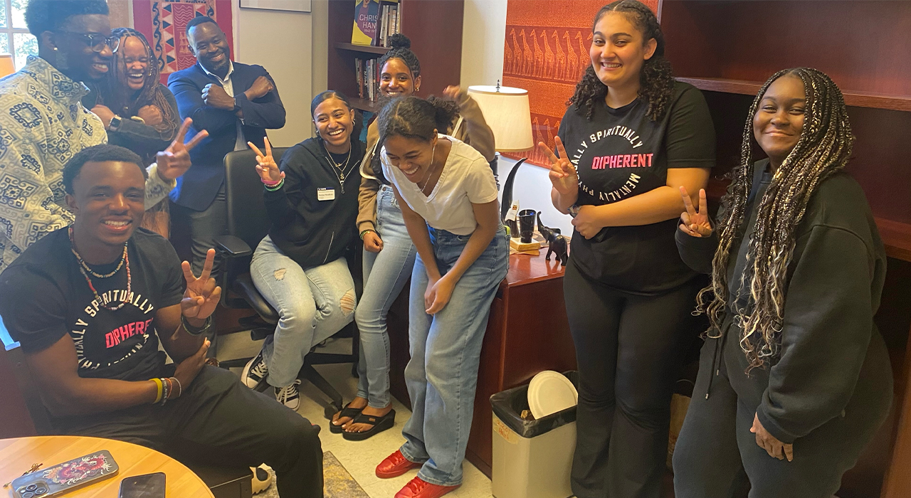 Eight students and their professor smile and laugh during office hours in the faculty member's office.
