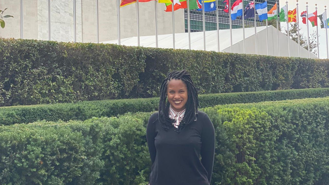 Andréia Coutinho Louback stands in front of a line of green hedges with different country flags flying in the background. She is outside on an overcast day. Andreia smiles and wears her black hair in long braids. She is dressed in a black sweater over a white and copper red striped collared shirt.