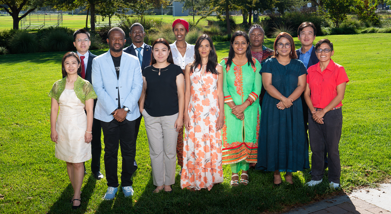 The 12 Humphrey Fellows stand posed in two rows outside on grass with an open lawn and bushes in the background.