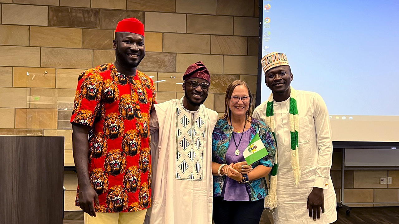 Collins wears a traditional red hat and red tunic with images of large roaring lion heads. He stands a head taller than the rest. Daniel wears a dark red traditional hat, glasses and a white tunic with blue embellishment on the front, Karen wears a colorful bright blue jacket with dark green and purple palm leaves over a purple top. She has glasses and a silver pendant on a black beaded necklace. Kurutsi wears a traditional white hat and tunic with a white and green blocked scarf. 