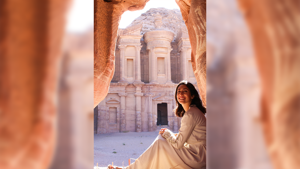 Victoria Casas smiles while sitting down on the floor with her legs pointed away from the camera. She wears her long brown hair down while wearing long sleeve white dress