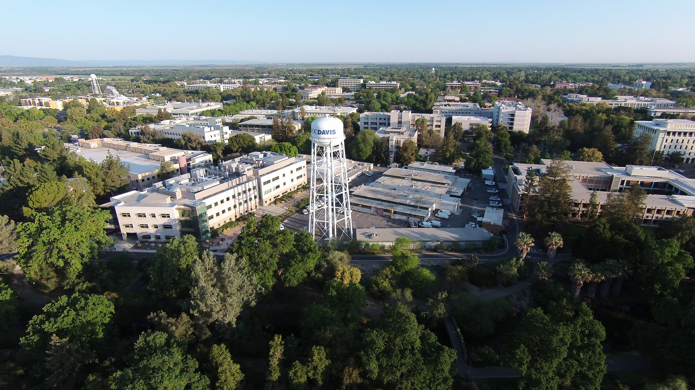 An aerial photo of the UC Davis Campus taken from the south and looking north. The photos shows the south water tower and Mrak Hall. It's a clear day with a blue sky, and trees and other campus buildings are spread out behind.  