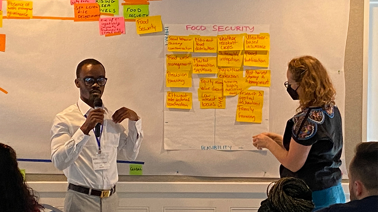 Sytekon “Caspar” Tarty holds a microphone as he stands in front of a white board with notes about the topic of climate change. He wears black glasses, a white collared shirt and khakis. His black hair is cropped short. Nikki Grey-Rutamu faces the board, affixing a note in place. Her strawberry blonde hair is shoulder length. She wears a black mask and black patterned top.