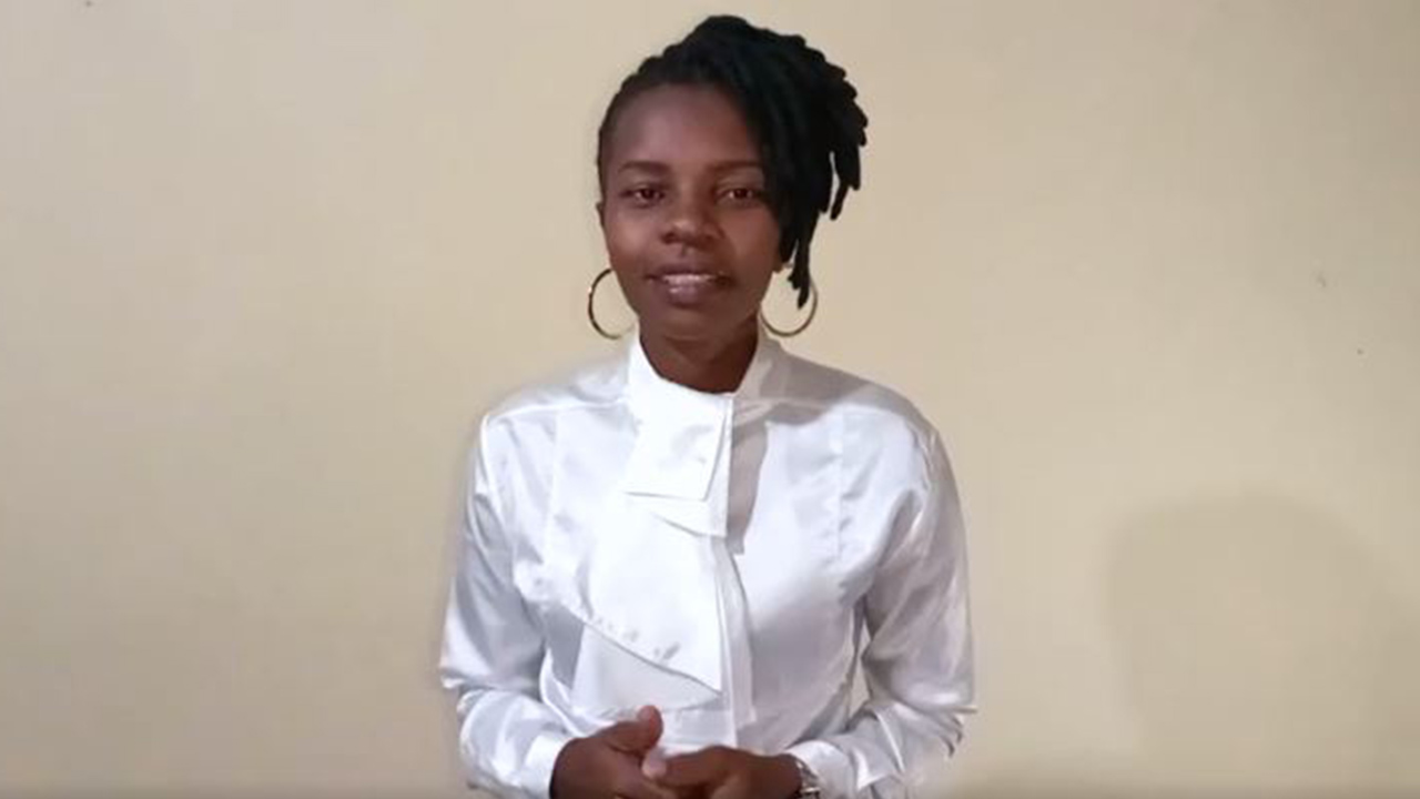 Esther Anyona stands before a white wall with her hands clasped in conversation. She smiles slightly and wears a white silk blouse with ruffles down the front, large gold hoop earrings, and her black hair is worn in locs pulled up to the top of her head and styled to her left.