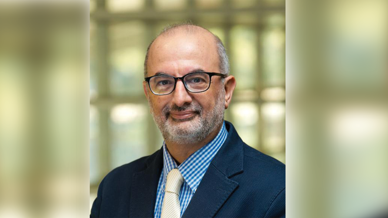 Fadi Fathallah smiles at the camera while wearing reading glasses, a navy blue suit jacket, light blue collared shirt, and yellow tie