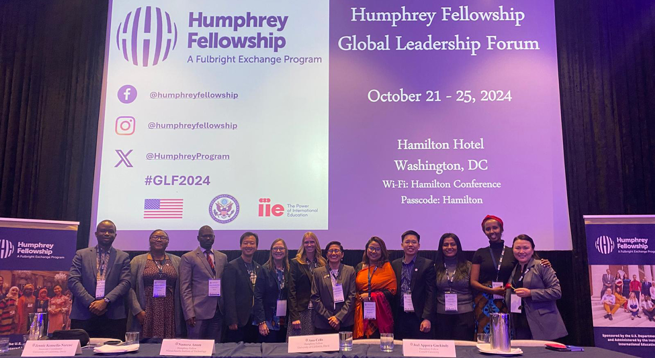 Twelve UC Davis Humphrey Fellows and two program coordinators stand in a single row behind a long table in front of signage that indicates they are at the Humphrey Fellowship Global Leadership Forum 2024.