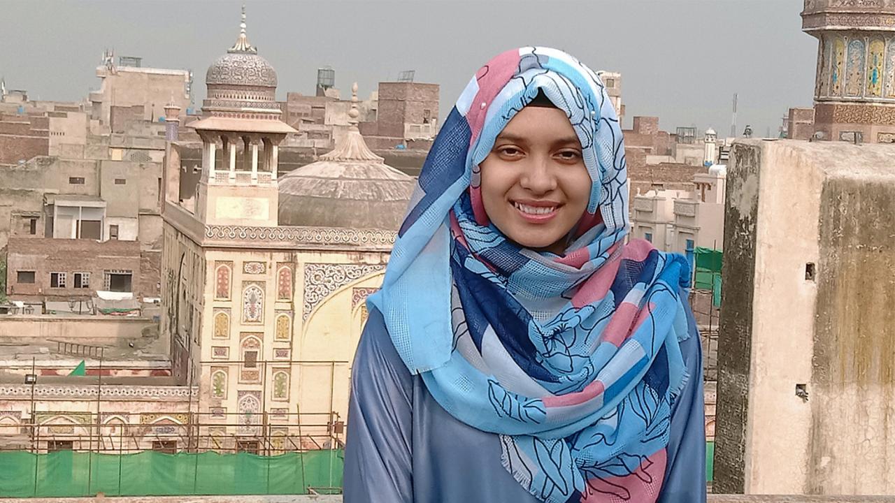 Hijab Khan smiles at a camera with a city in Pakistan behind her. She wears a blue shirt and a blue and pink headscarf. 