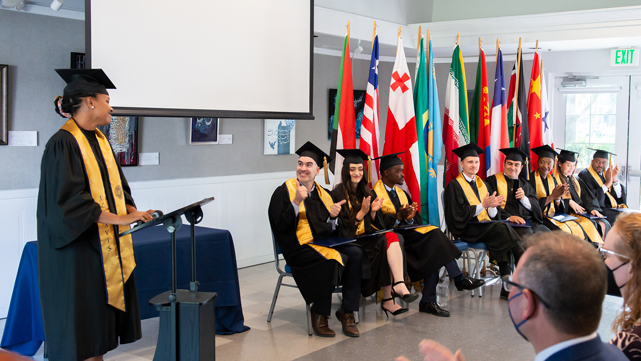 Andreia Coutinho Louback stands in graduation robes at a podium and addresses the other Humphrey fellows seated in a row next to her, also dressed in graduation robes. 