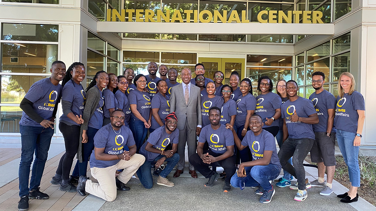 UC Davis Chancellor Gary S. May stands in the center of a group of Mandela Fellows wearing a light gray suit and smiling at the camera. Surrounding him are 25 Mandela Washington Fellows and two program coordinators, all wearing blue T-shirts that have a circle logo and UC Davis Global Affairs on the front.