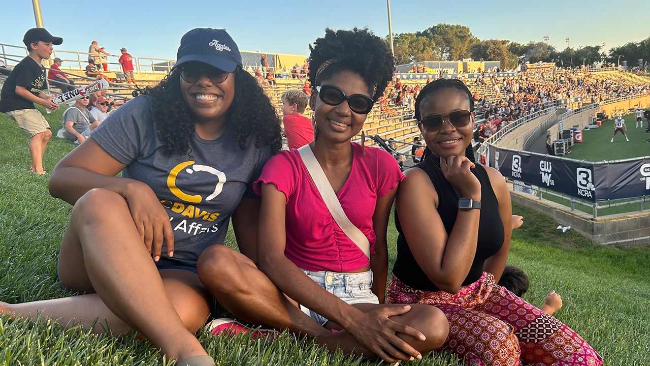 Kara sits on the left and wears a blue baseball cap that says "Aggies" in a white script font on the side and a Global Affairs T shirt. Rosani sits in the middle and Mpho on the right. They sit on the grass with the stands, spectators and sideline of the football stadium behind them. 
