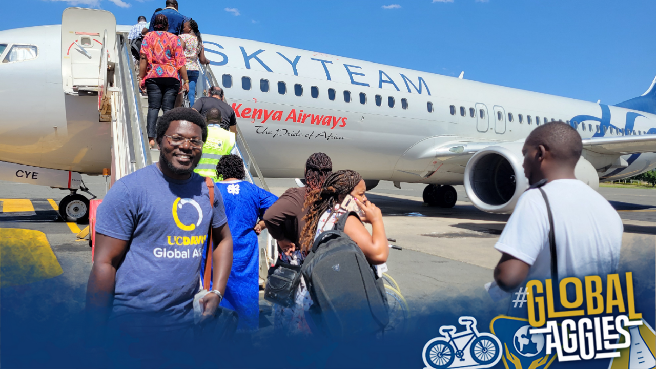 Roy Akumu smiles while wearing a UC Davis Global Affairs shirt in front of a Kenya Airways plane