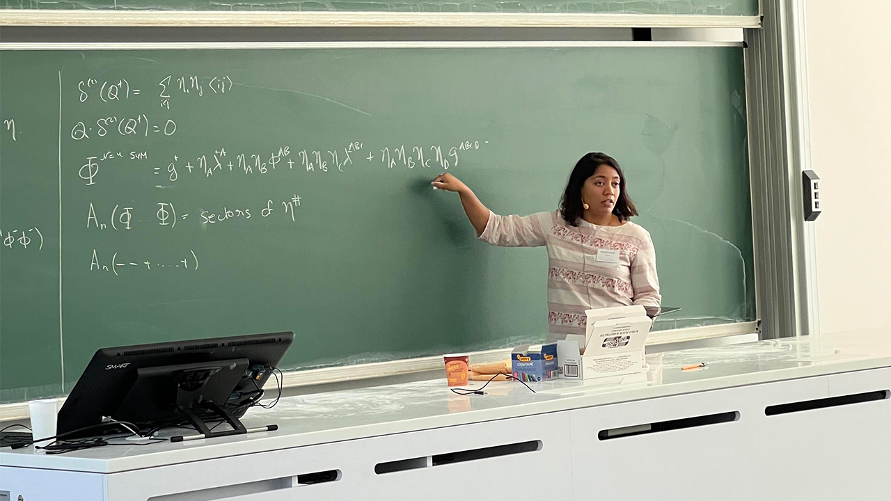 Shruti stands behind a white platform in a lecture hall, gesturing to the green chalkboard directly behind her at one of several equations written there. She is speaking to a crowd off camera.