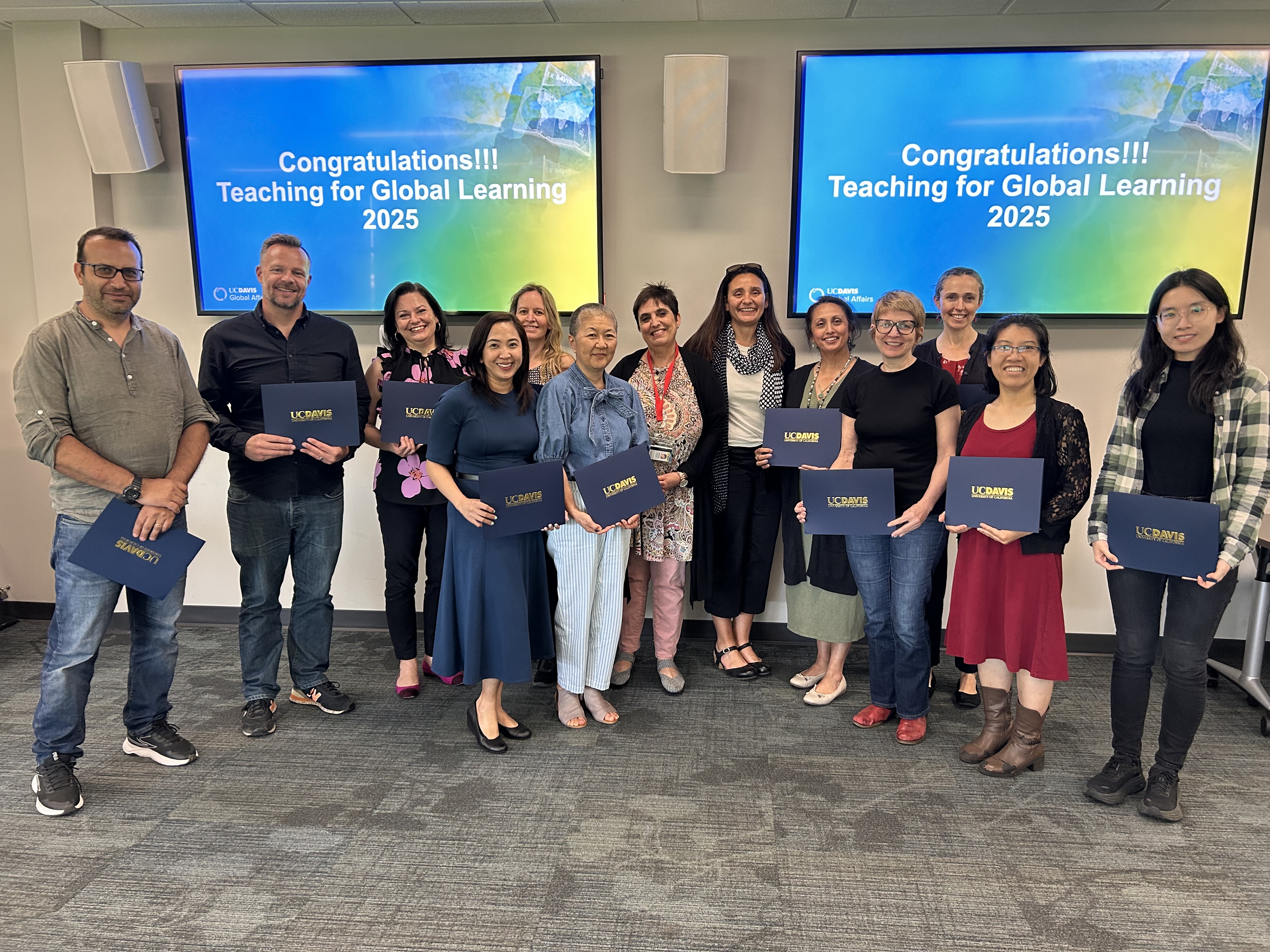 A group of UC Davis faculty members smile at the camera with Congratulations! Teaching for Global Learning 2025 on the two screens behind them. 