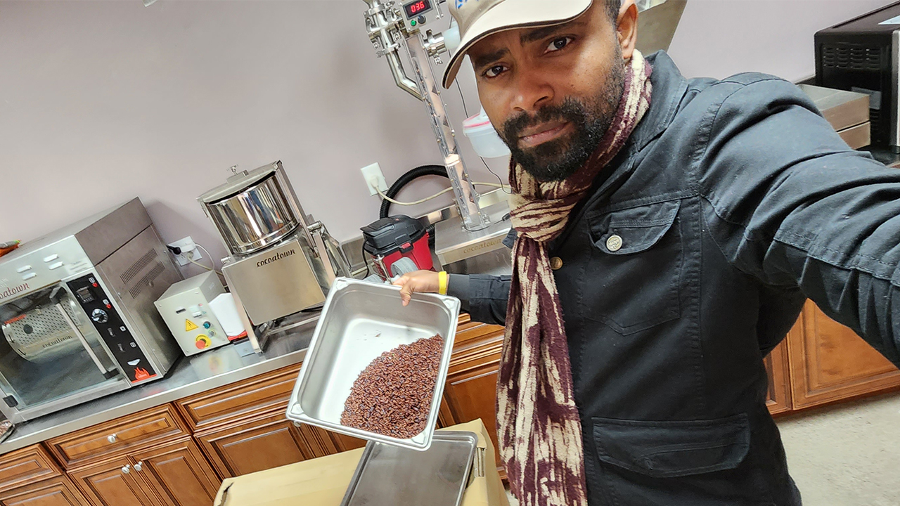 Edu takes a selfie of himself in a kitchen lab setting as he holds a stainless steel container of cocoa beans.