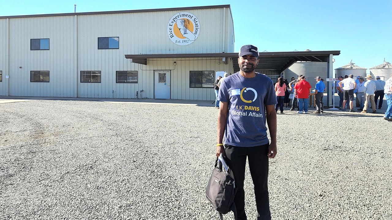 Edu wears a UC Davis Global Affairs T-shirt and stands outside the main California Rice Experience building. The building, a gathering of people and California Rice Experiment signage are in the background. 