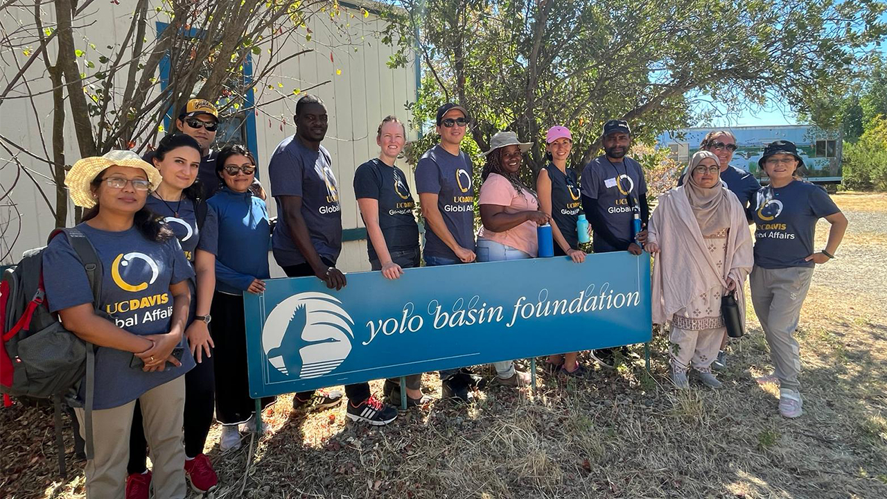 Sushila and 12 members of the UC Davis Humphrey Fellowship stand outside around a blue sign that says "Yolo Basin Foundation" in white. Most wear UC Davis Global Affairs T-shirts and are dressed for the sunny weather. 