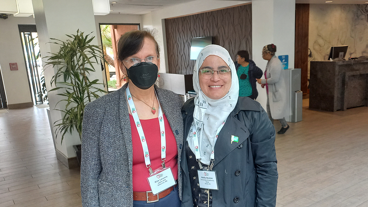 Nadia and Dr. Lewers stand in a lobby. Dr. Lewers wears a black facemask and both women are dressed in gray blazers with their conference badges hanging from lanyards around their necks. 