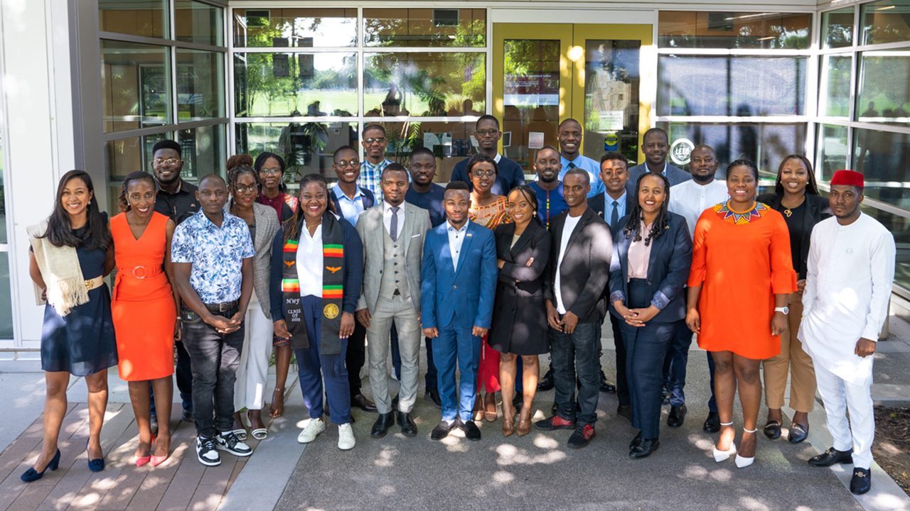 A group of 25 Mandela Washington Fellows stand in front of the glass windows of the International Center in the shade.