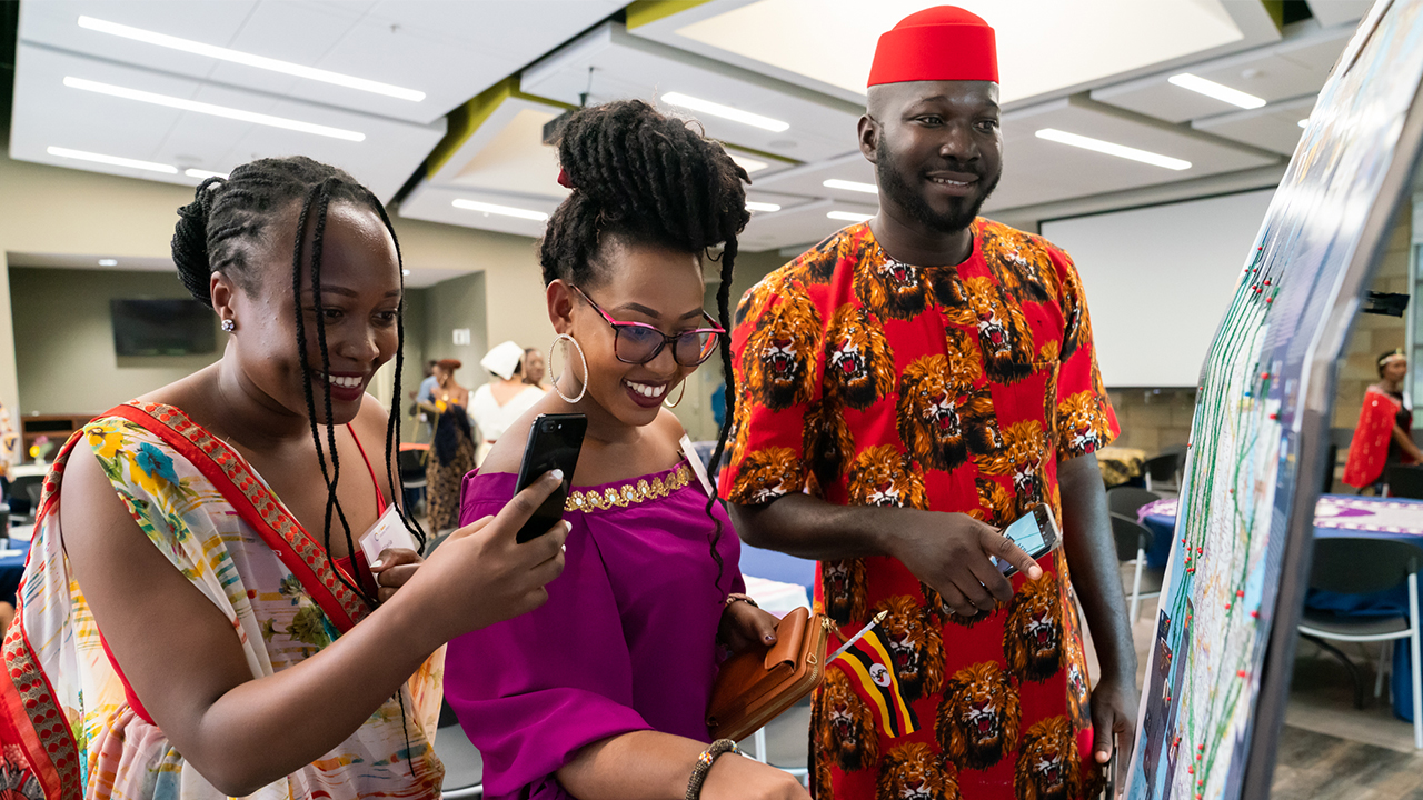 Three fellows in traditional outfits stand at a poster map on an easel. They all smile toward the map as Gloria is poised to take a photo of it with her phone and Gloria appears to be gesturing just out of frame. 