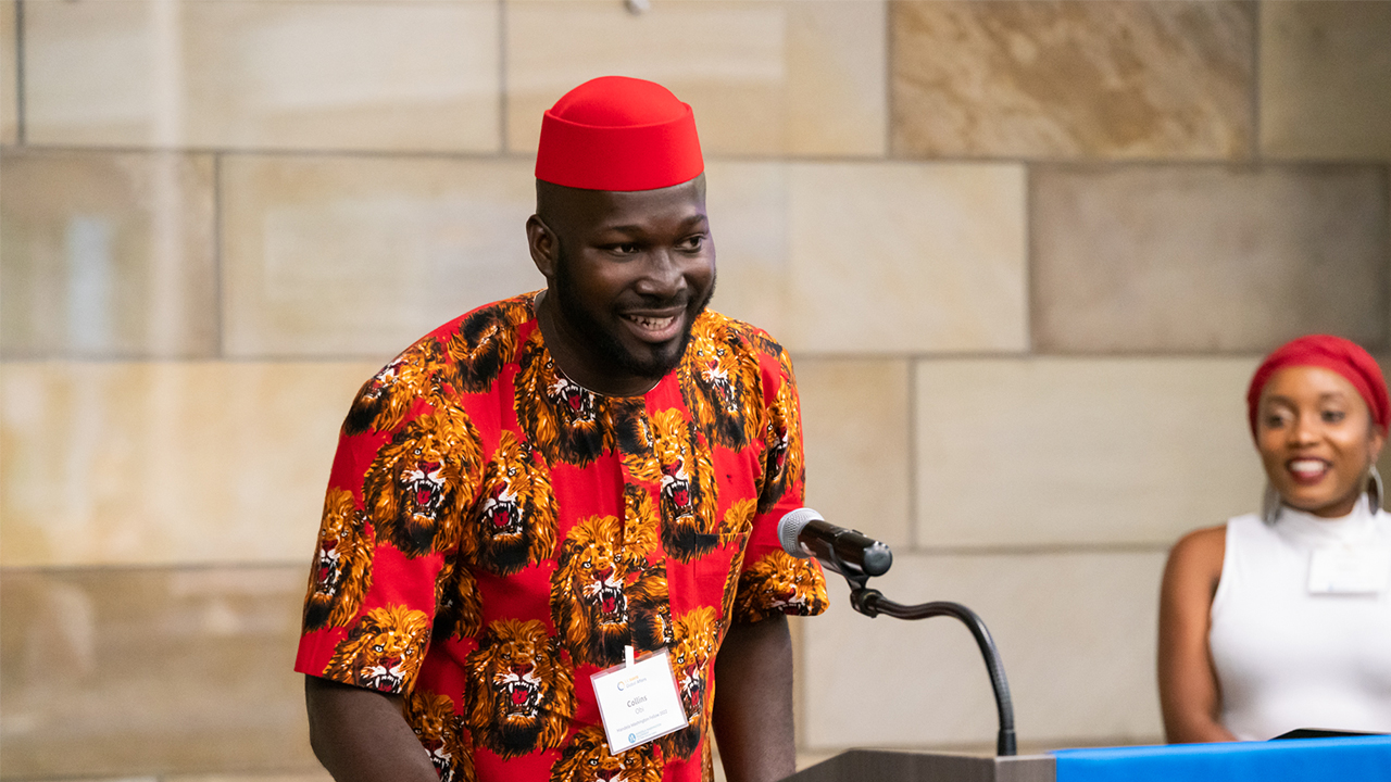 Collins speaks at a podium. He wears a red hat and a red tunic with large roaring lion heads. 