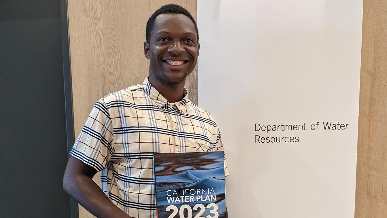 Immanuel smiles next to a white wall panel that reads in basic black lettering "Department of Water Resources". He wears a cream short sleeved collared shirt with a navy, light blue and red plaid. He holds a folder with a water background that reads in white block lettering, "California Water Plan 2023."