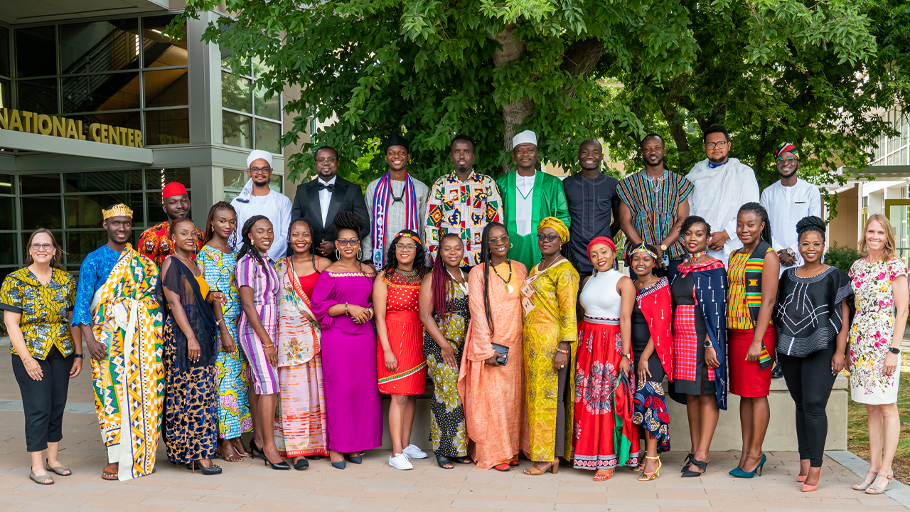 25 Mandela Washington Fellows and two administrators stand in two rows under a large tree outside the International Center to the left of the photo. The fellows all wear colorful traditional dress from their African nations.