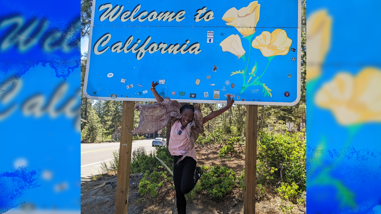 Marilyn jumps with her hands in the air and smiles in front of a blue sign with yellow poppies that reads "Welcome to California"