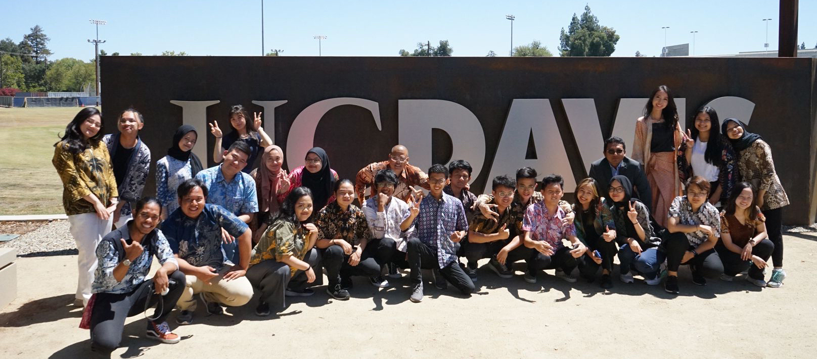 The group of students from Indonesia and members of the Office of the Consulate General squat, kneel and stand in front of a large gray stone wall with large white letters that read "UC Davis". This sign on the edge of campus is in the sunlight on a warm and sunny day.