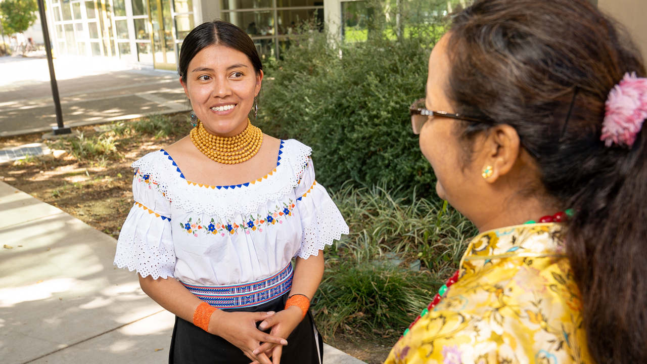 Tamia wears a traditional Ecuadorian white blouse with rows of yellow beads adorning her neck. She smiles at Sushila, who is visible from behind, wearing a yellow floral top. The women stand outside of the International Center.