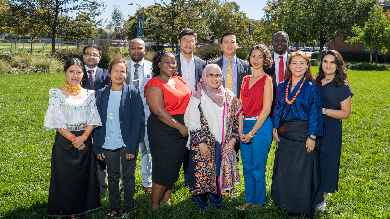 Standing in two rows with men in the back row and women in the front, the twelve Humphrey fellows from the 2022-23 cohort are smiling and looking at the camera. They wear various business professional and customary ethnic dress. They are on grass in front of the International Center with the campus behind them and a blue and yellow UC Davis banner hangs on a light pole in the distance.