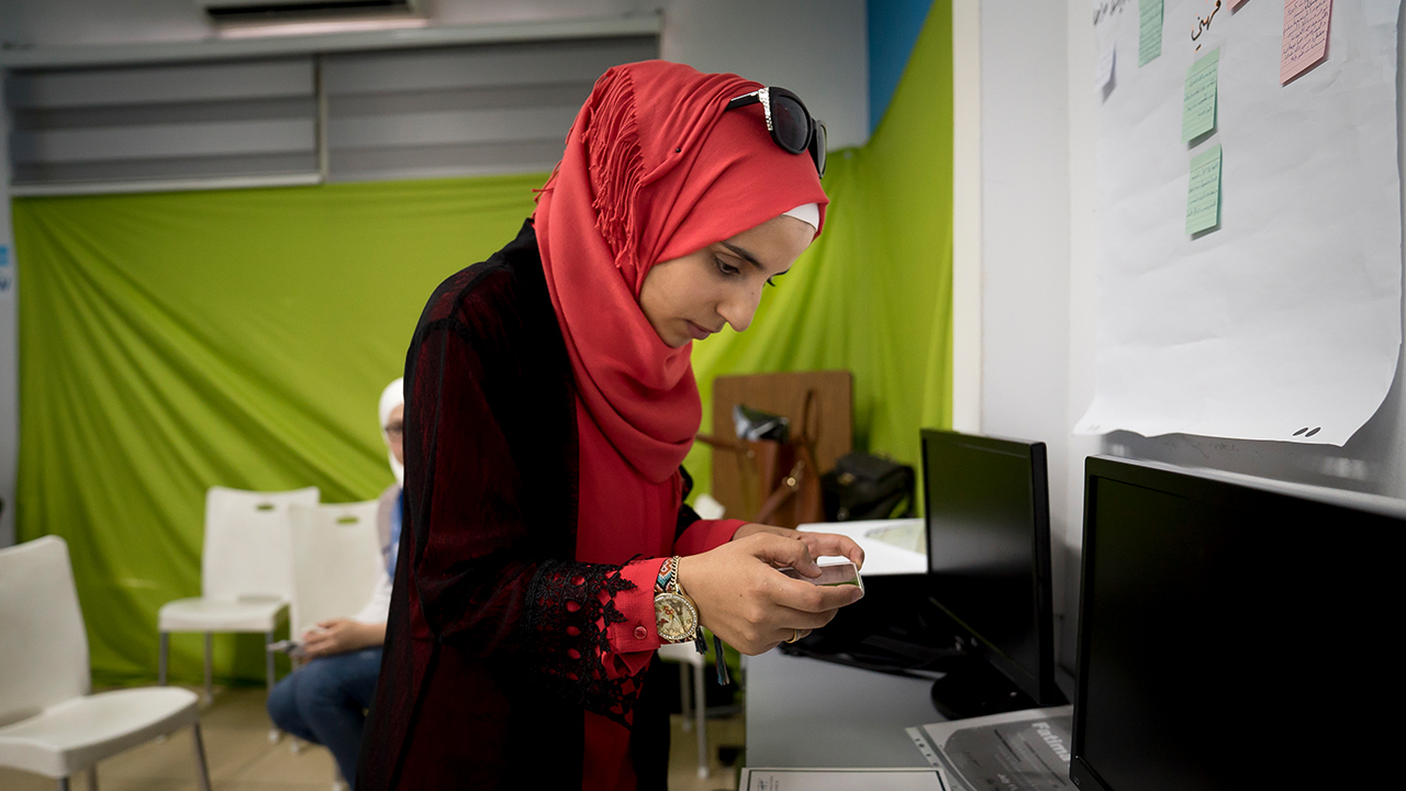 A woman in a red hajib and black top stands to look over paperwork in an office. There is a white board and a bright green wall behind her.