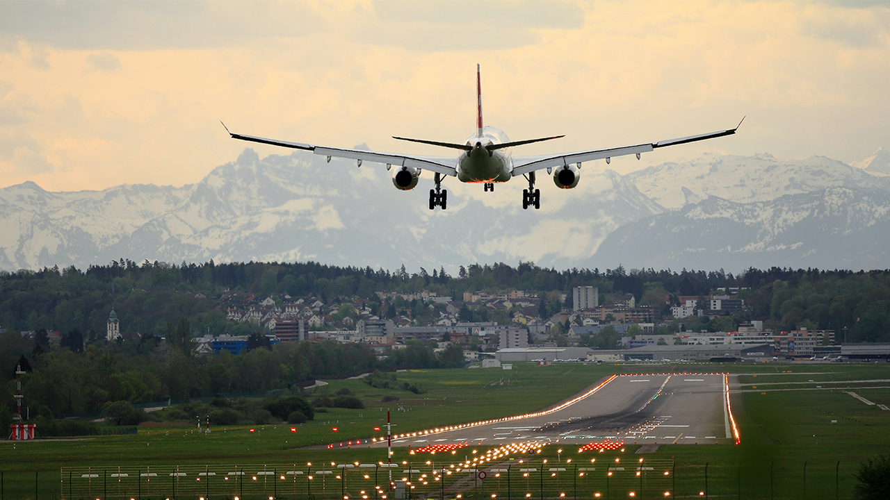 An airplane descends toward a runway at an airport in Zurich. The city and Swiss mountains unfold in the background. 