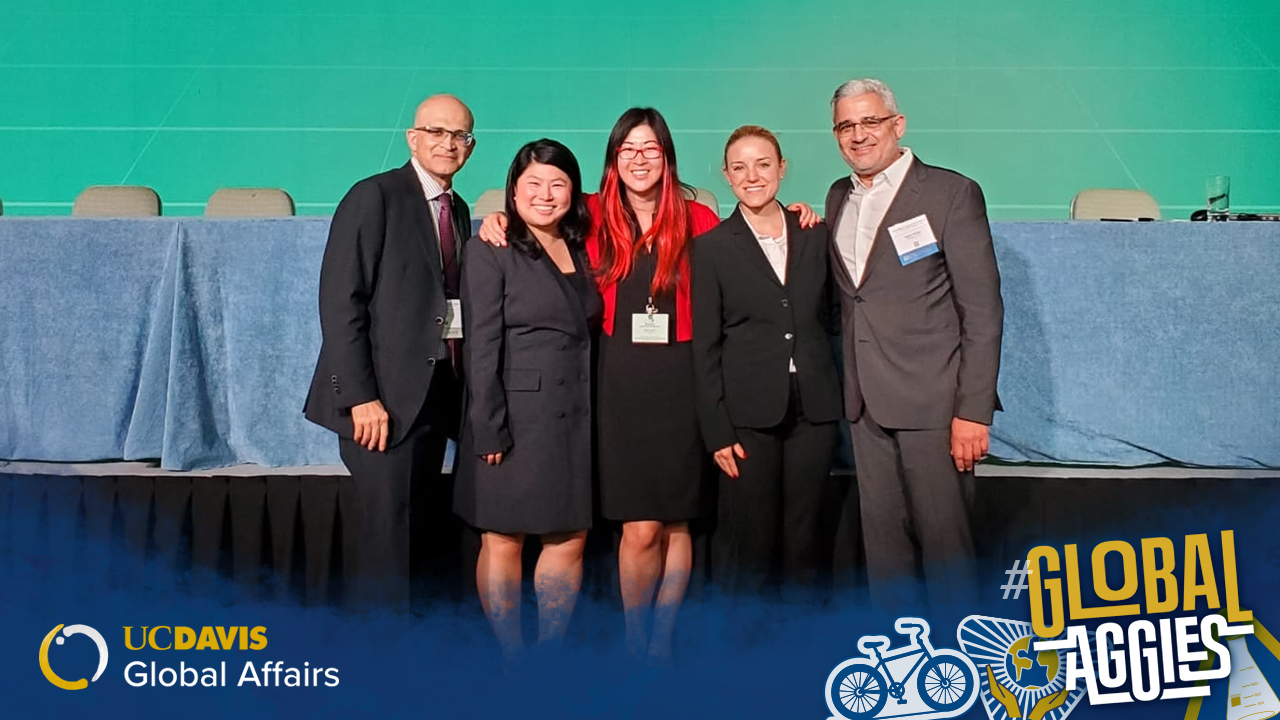 Bob Kiaii stands in front of a stage with three woman and one man all wearing business attire. The stage behind them is set up for a panel of speakers with draped tables and chairs in front of a large light green screen. A graphic with the UC Davis Global Affairs logo and #GlobalAggies lays over the photo at the bottom.