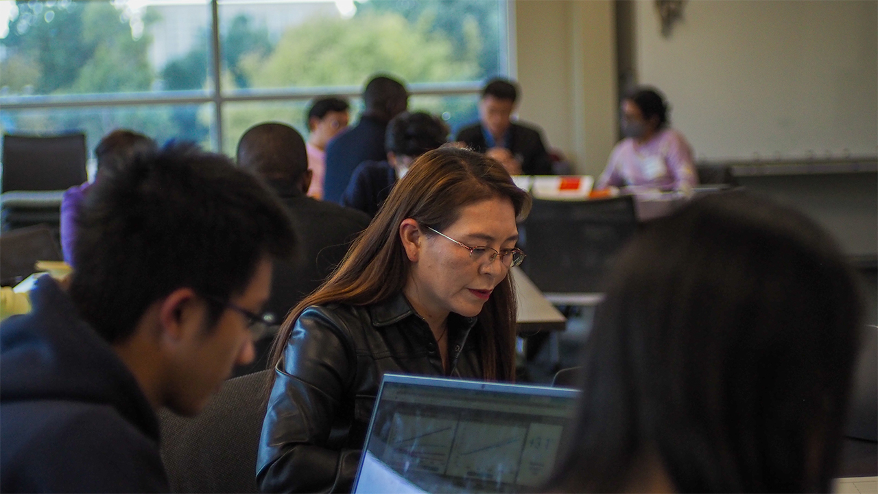 Samtso wears wire rimmed glasses as she looks at her computer screen. Behind her in the room are other negotiation groups. In the foreground, we see over the shoulder of a student who is looking at the graphs and charts within the simulation tool. 