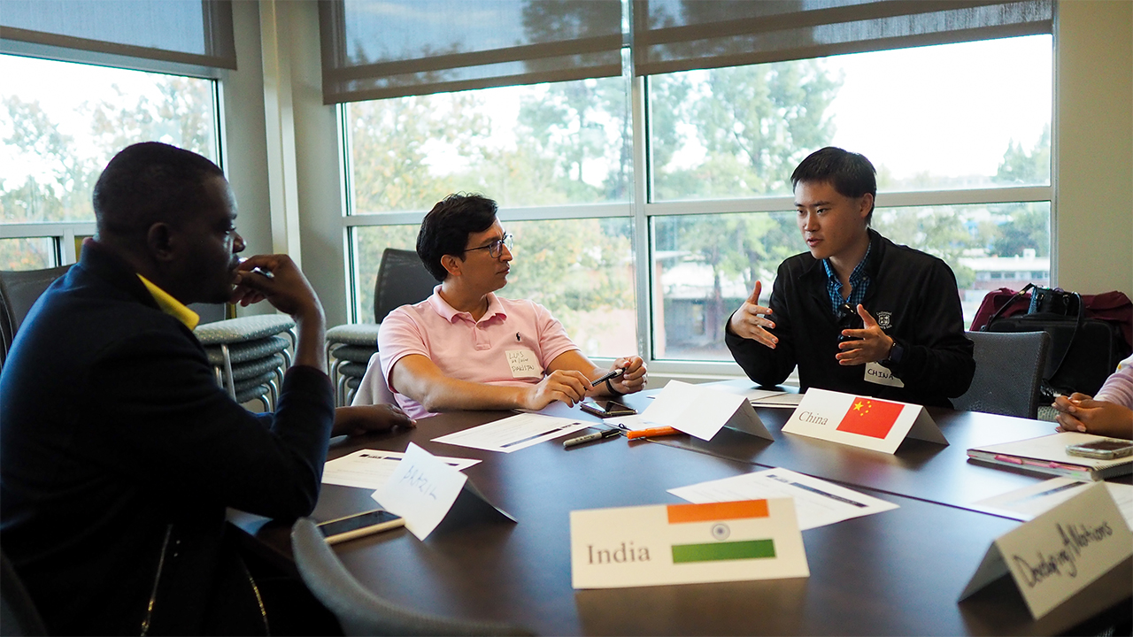 Silas sits in profile at a work table with Luis at his left. They both look at and listen to a male student representing China at the right of the photo, who is speaking and gesturing conversationally with his hands. Placards on their table identify the group as "Developing A Nations" and participants represent countries including China, India and Brazil.