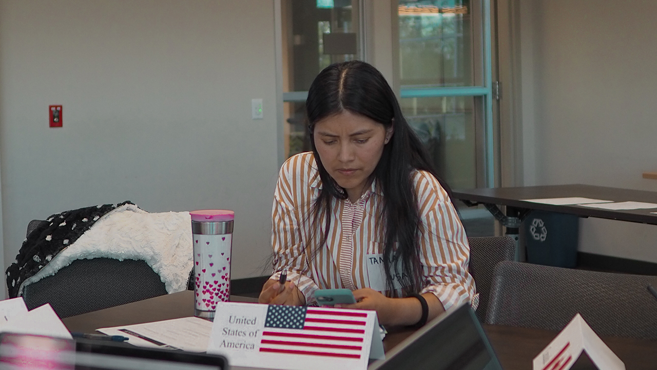 Tamia sits at a work table and looks at notes on her mobile device during the climate change simulation exercise. The placard in front of her identifies her as representing the United States of America.