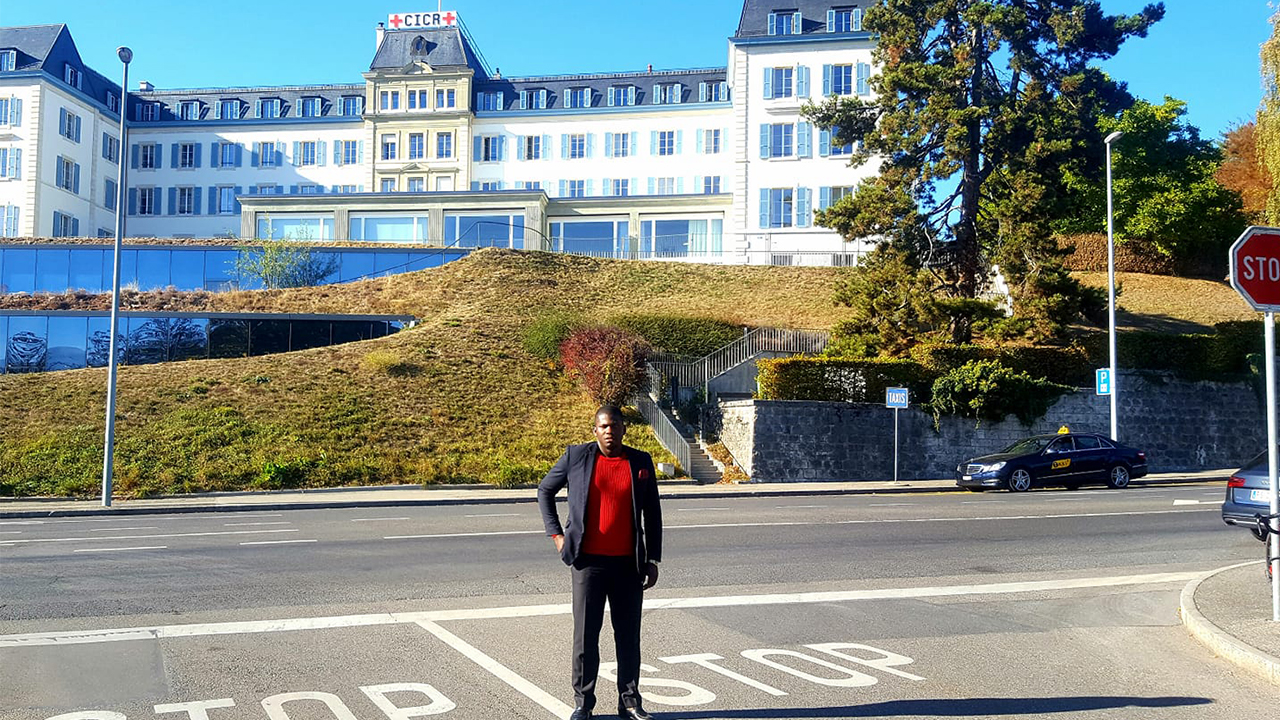 Jireh Bookwa stands at a stop sign at an empty three-way intersection in front of the Red Cross building in Switzerland, which sits elevated on a hill. It is a sunny day, and Jireh wears a red sweater under his dark gray suit. His right hand is in his jacket pocket. His black hair is closely cropped. Behind him, the white building stands five stories tall and modern additions of two levels of glass windows have been built into the hill below. 