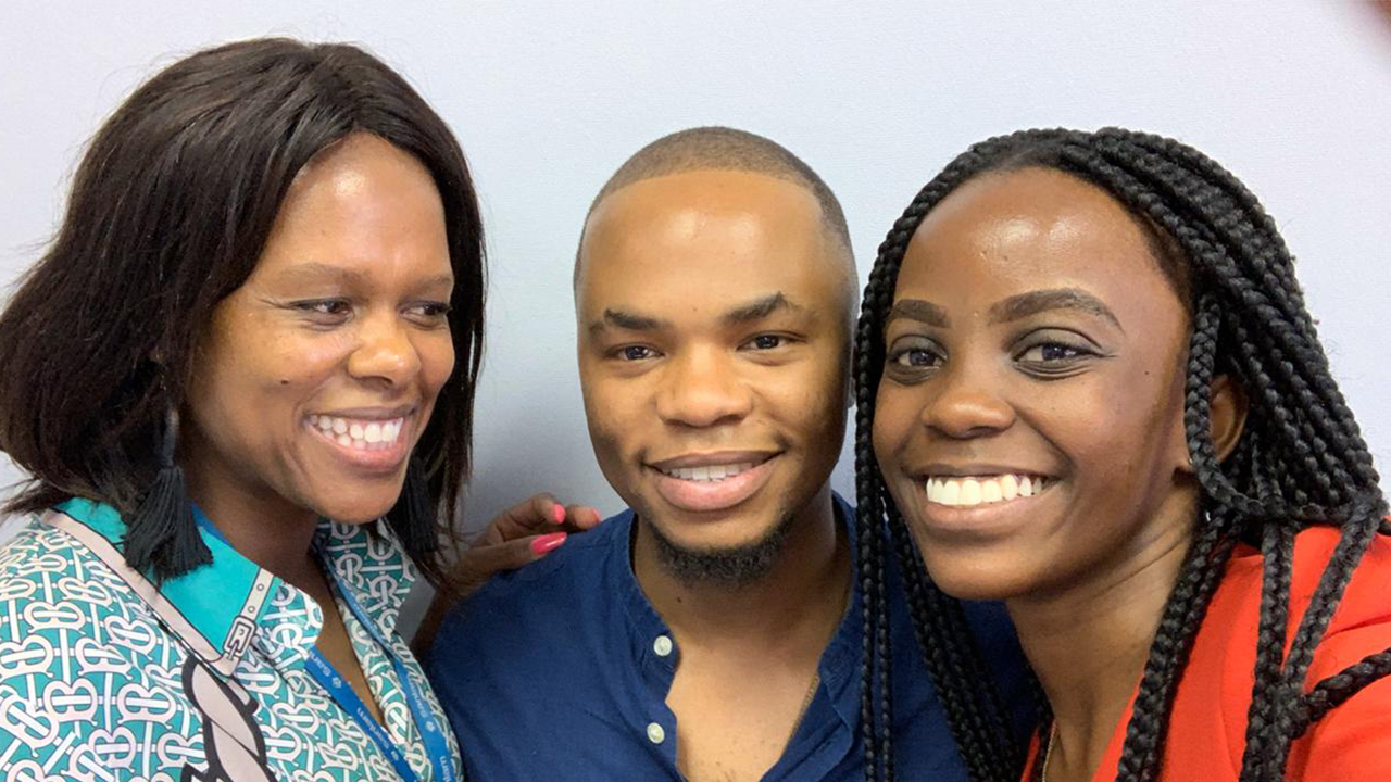 Kwezi Sogoni stands between two women smiling for the camera. He wears a dark blue collar-less button-down shirt, his black hair is cropped close and he has a small beard on his chin. His supervisor, Mamatlhare Maseko is on his left. She wears black tassel earrings, a turquoise patterned blouse, and her black hair is work straight in a bob. On is right, Mbali Motsoaneng wears a red blazer and her long black hair is worn in braids. 