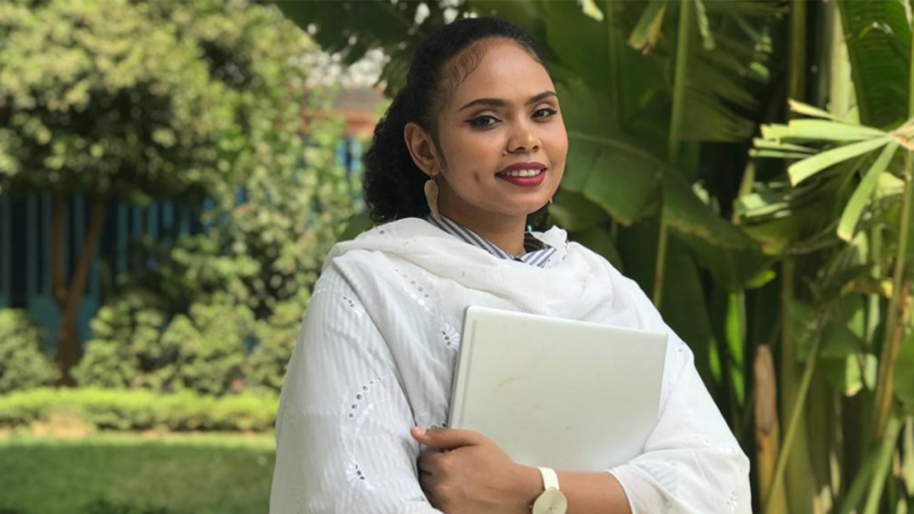 Samia Said stands outside in the shade on a sunny day in front of trees and palms. She wears a gray and white striped collared shirt covered by a draped white robe, clutches a white laptop to her chest and wears a large gold watch on her left wrist. Samia smiles at the camera. Her black curly hair is pulled back into a ponytail and she wears large gold earrings in the shape of wings or feathers.