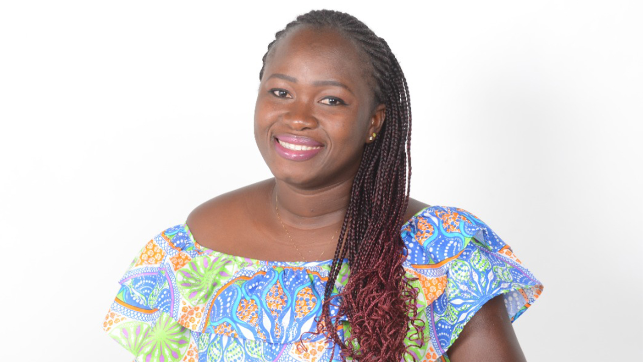 Titchinmin Kone sits before a white background. She wears a colorful pastel patterned off-the-shoulder dress with lavender, aqua, orange, and bright green. She smiles broadly at the camera. Her dark hair is worn in braids with dark pink ends, and she wears a gold chain necklace and two stud earrings in white and green.