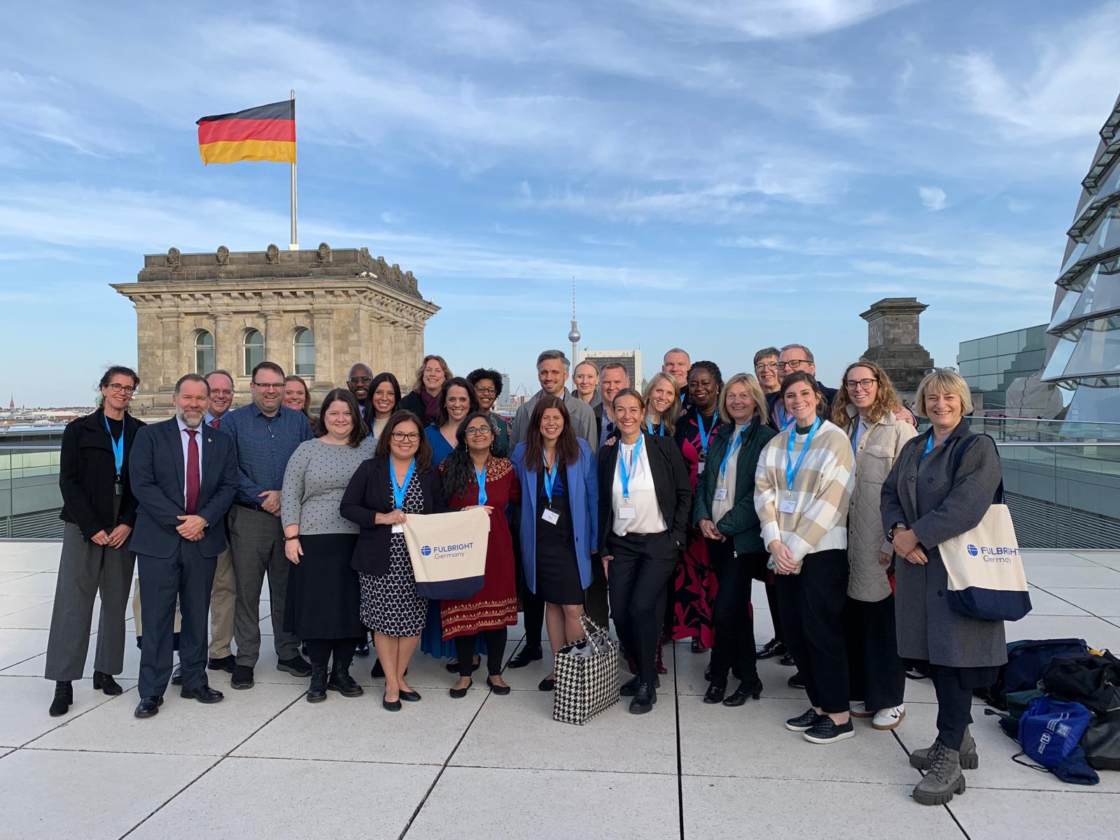 A group photo of Fulbright participants on the Reichstag rooftop in Berlin. The German flag waves atop the building, and the photo captures a mix of historic architecture and modern skyline elements, including the Fernsehturm tower.