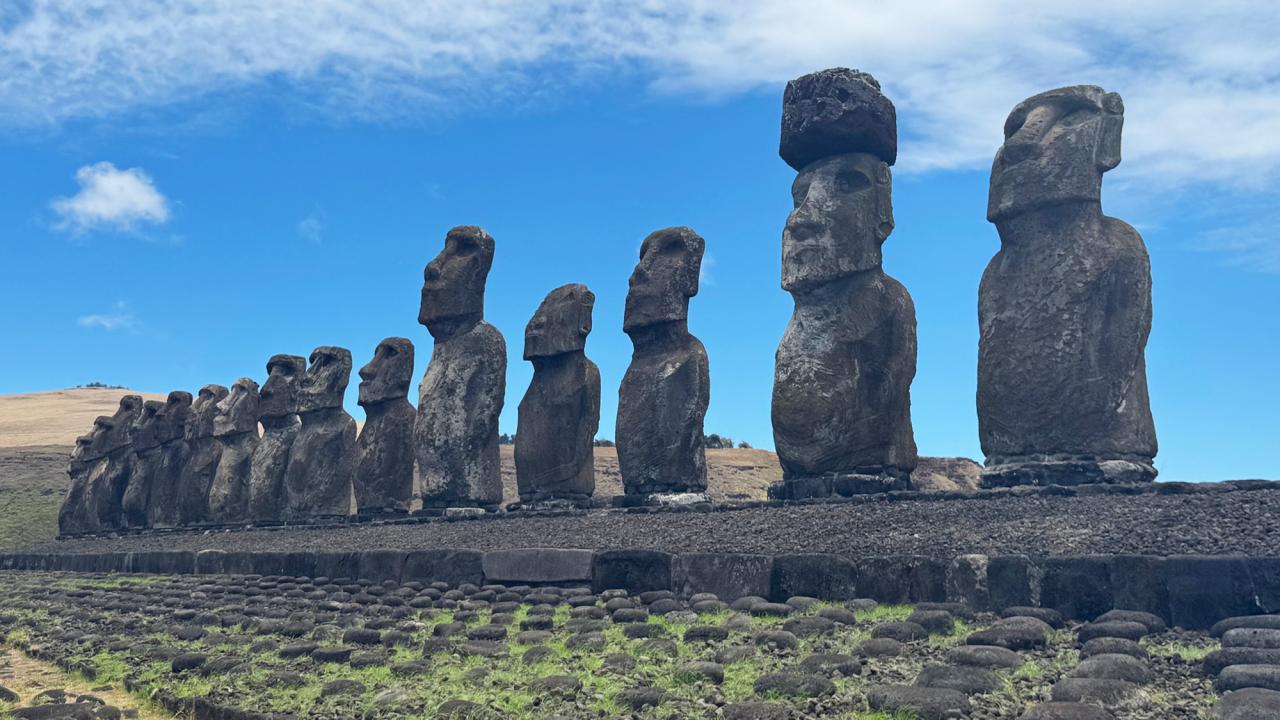 Row of famous statues on Easter Island with hills in the background and a grassy stone foreground.