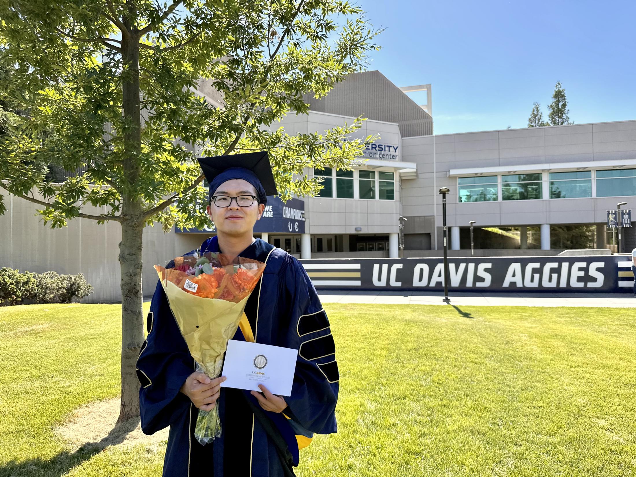 Jeff Lai holding flowers and his diploma, standing in front of a large UC Davis Aggies sign.