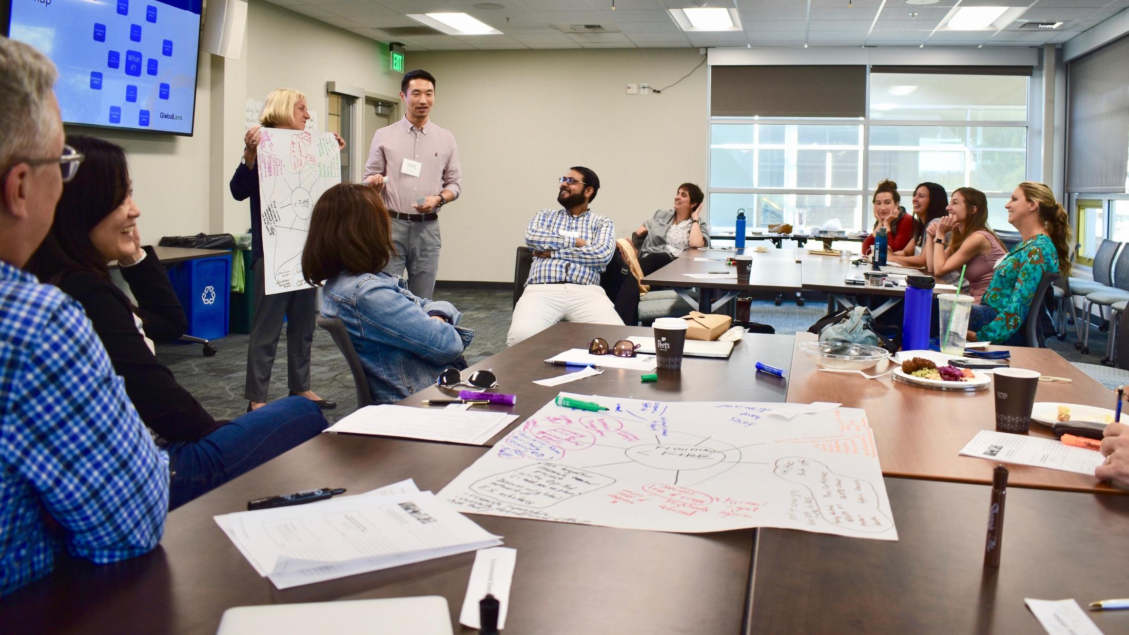 A group of people working at a table look at a presenter holding up a large paper