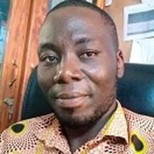 Komi Edem Assigbley sits in a tall black office chair with a cork bulletin board behind him wearing a tan collared shirt with an abstract circular pattern in black, yellow and white. His dark hair is shortly cropped and he smiles slightly at the camera. 