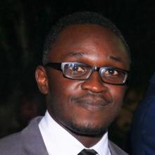 Headshot of Bertin Ciza taken at night with a dark background as he stands in the crisp light. He wears a gray suit jacket over a white collared shirt, black striped tie, and dark-rimmed glasses. His black hair is short, and he has a slight smile as he looks at the camera